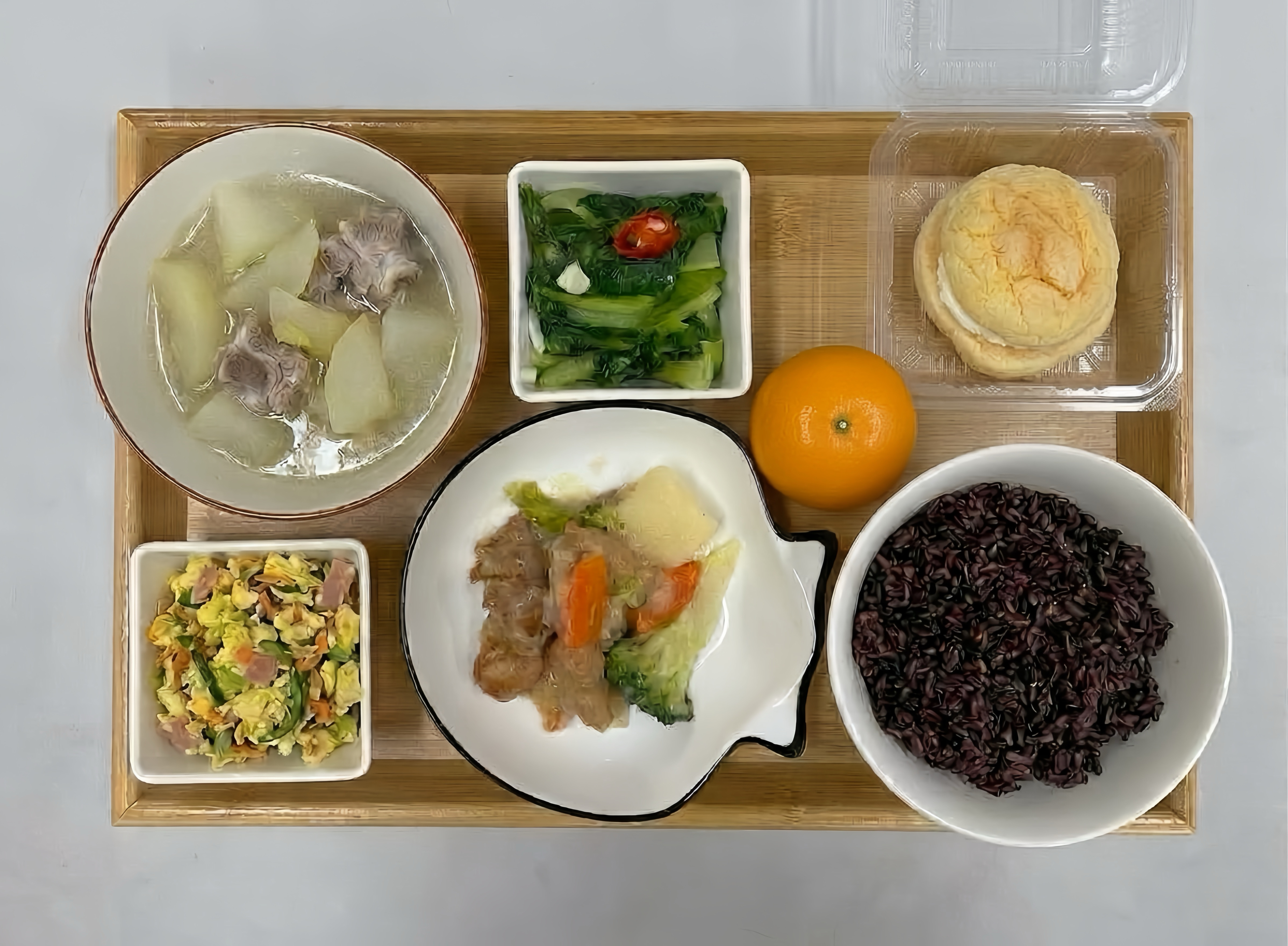 Actual Week 6 school lunch tray with fish in cream sauce, carrots and broccoli in a shell-shaped dish, purple rice, soup, green vegetables, an egg dish, an orange and a bun